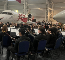 A live orchestra performance inside an aircraft hangar, with musicians dressed in black playing various instruments. A conductor leads the ensemble, and an airplane is visible in the background. The event is part of the 'Veterans Through Music' program.
