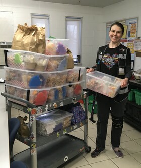 Smiling woman standing in a kitchen holding a plastic container filled with snacks, next to a cart stacked with bins of pre-packaged food items. Community food program, with a focus on providing nutritious meals