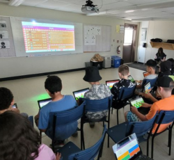 A classroom of students engaged in an interactive French learning session using laptops. A large screen at the front displays a colorful game leaderboard, indicating a gamified approach to language learning. The students, seated in rows, are focused on th