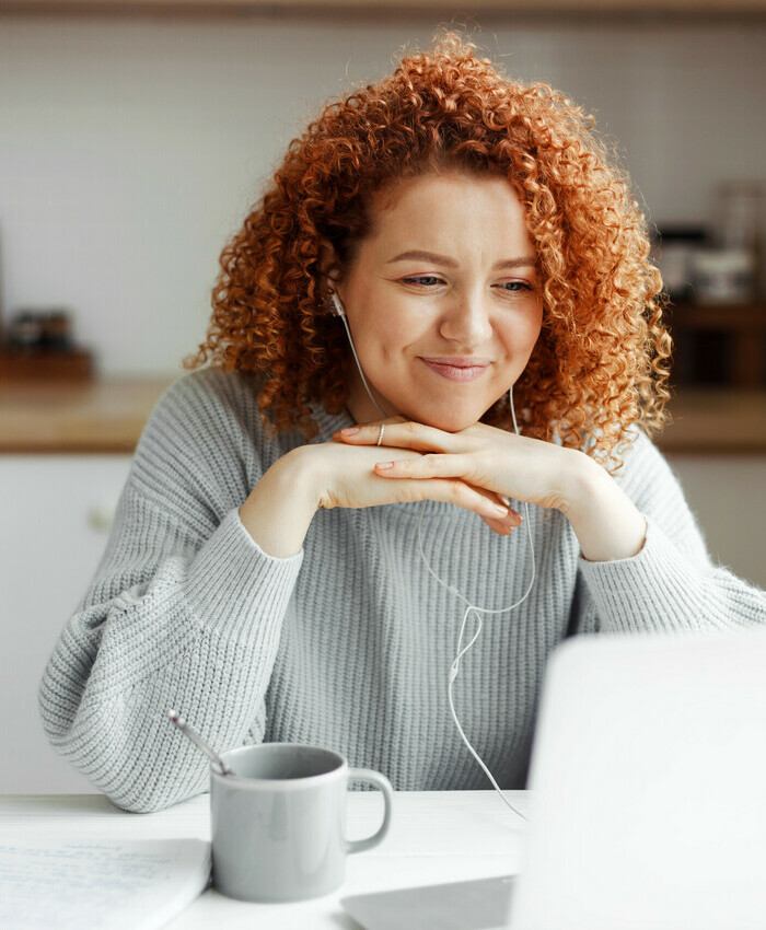 Smiling parent sitting at kitchen table with coffee cup and pen and paper, watching a webinar on their laptop with headphones in